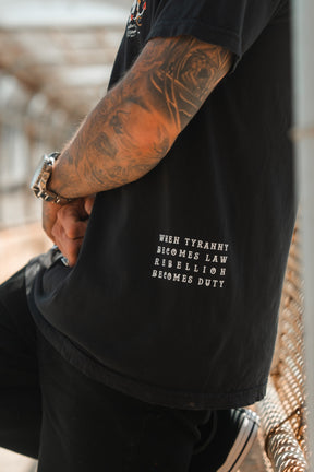 A tattooed person sits sideways on an outdoor bench wearing the Relentless Betrayal Revolution Premium Tee in black, featuring white "WHEN TYRANNY BECOMES LAW REBELLION BECOMES DUTY" text.