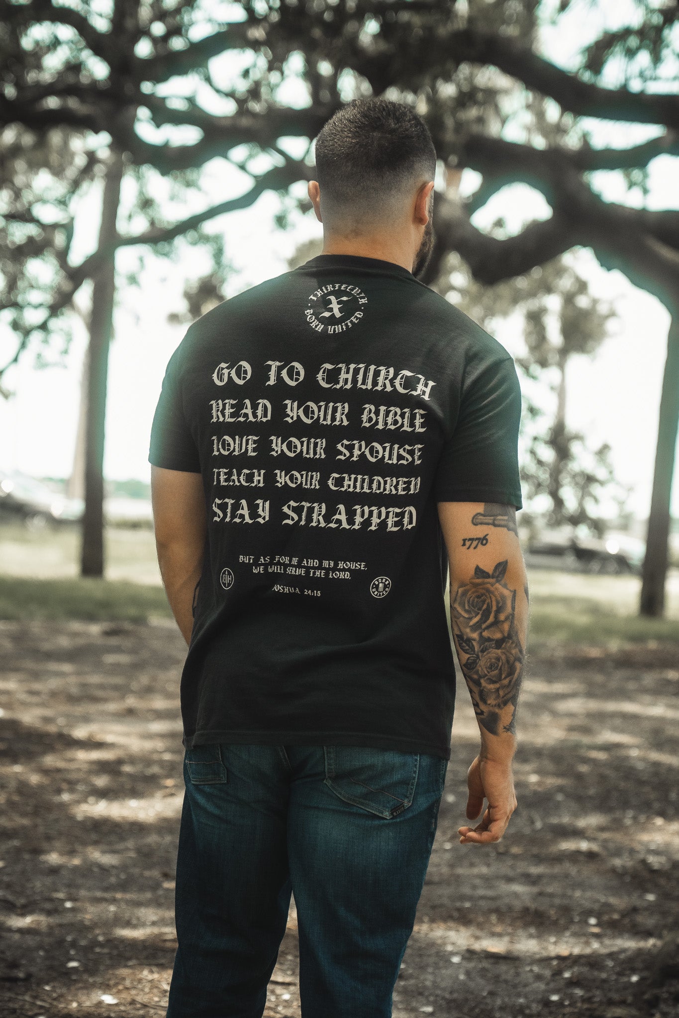 A man stands outdoors with his back to the camera, wearing the Kingdom Protector Premium Tee by Born United Private Label, featuring "Go to church, read your Bible, love your spouse, teach your children, stay strapped" and forest trees in the background.