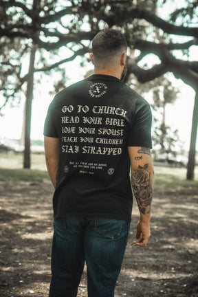 A man stands outdoors with his back to the camera, wearing the Kingdom Protector Premium Tee by Born United Private Label, featuring "Go to church, read your Bible, love your spouse, teach your children, stay strapped" and forest trees in the background.