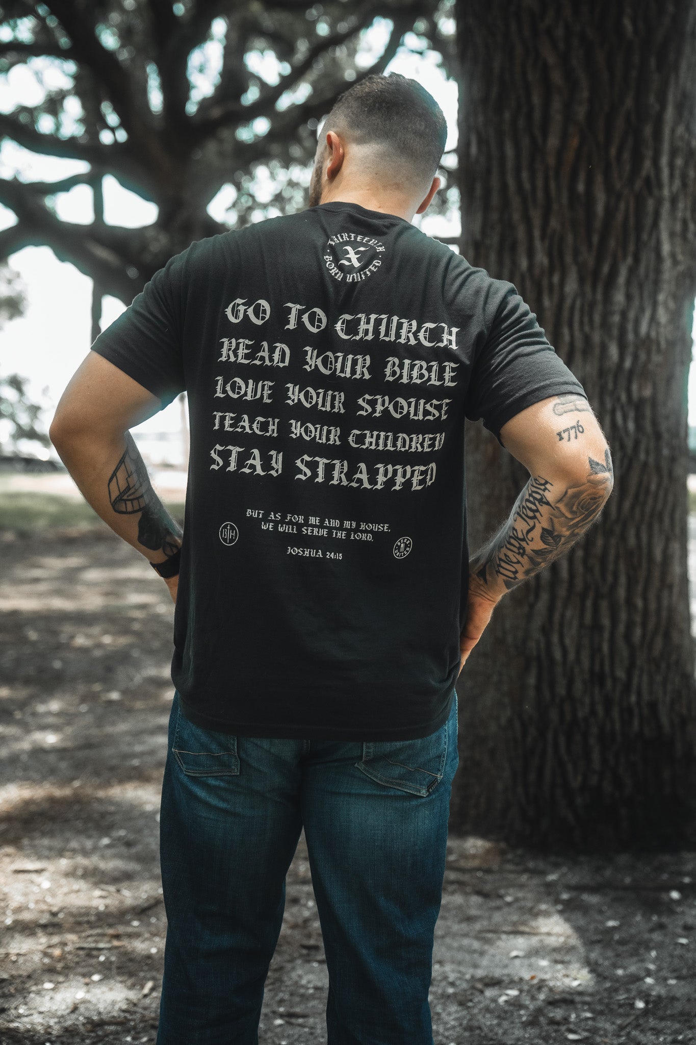 A man stands outdoors with his back to the camera, wearing the Kingdom Protector Premium Tee by Born United Private Label featuring: Go to church, read your Bible, love your spouse, teach your children, stay strapped—values from Joshua 24:15.