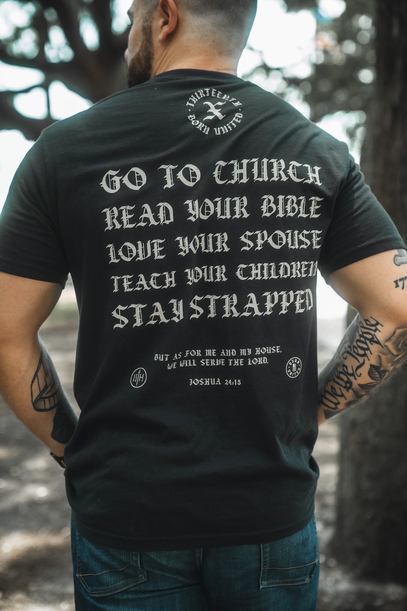 A person stands outdoors in a Born United Private Label Kingdom Protector Premium Tee featuring “GO TO CHURCH, READ YOUR BIBLE, LOVE YOUR SPOUSE, TEACH YOUR CHILDREN, STAY STRAPPED" with Joshua 24:15—ideal for Christian home decor or wall art inspiration.