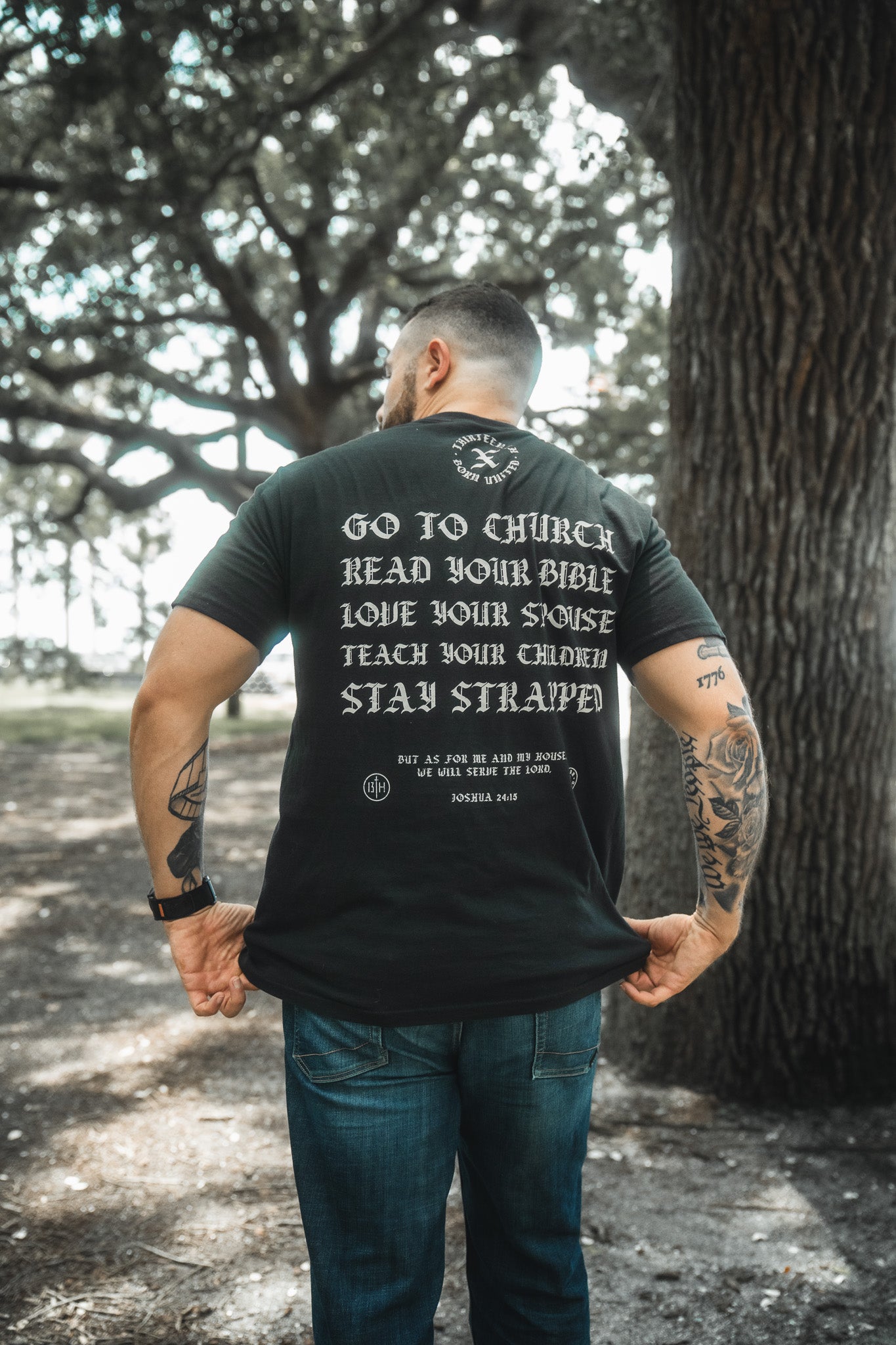 A tattooed man stands outdoors, facing away, wearing the Kingdom Protector Premium Tee by Born United Private Label. The black t-shirt displays "Go to church, read your Bible, love your spouse, teach your children, stay strapped," referencing Joshua 24:15.
