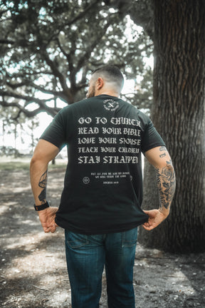 A tattooed man stands outdoors, facing away, wearing the Kingdom Protector Premium Tee by Born United Private Label. The black t-shirt displays "Go to church, read your Bible, love your spouse, teach your children, stay strapped," referencing Joshua 24:15.