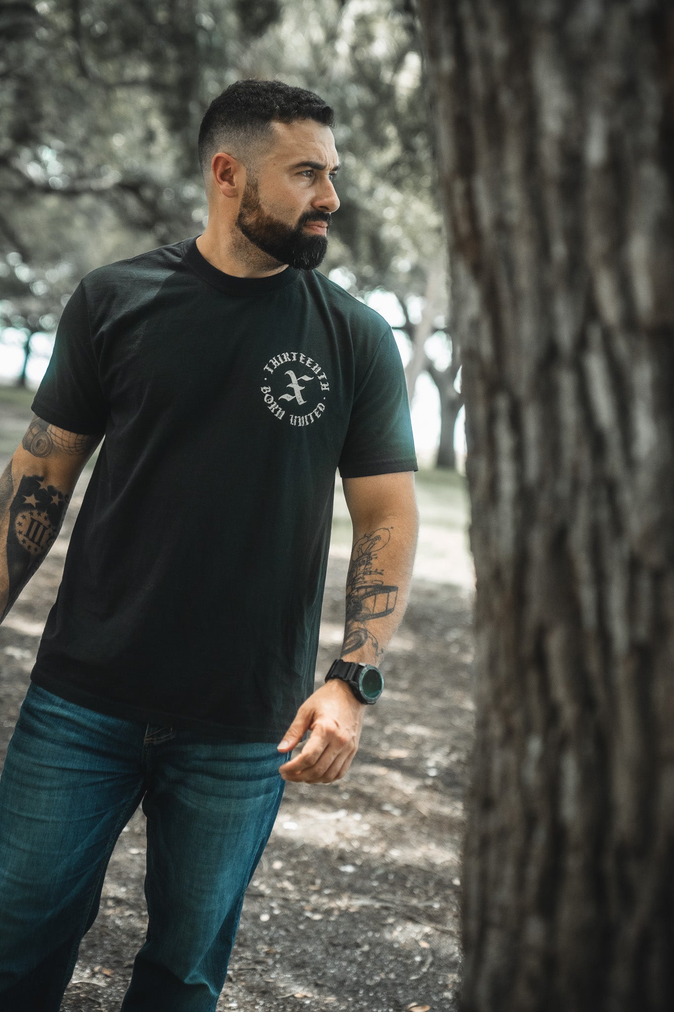 A bearded, tattooed man wearing the Kingdom Protector Premium Tee by Born United Private Label stands outdoors near a tree, with blurred greenery in the background evoking Christian serenity like Joshua 24:15 wall art.