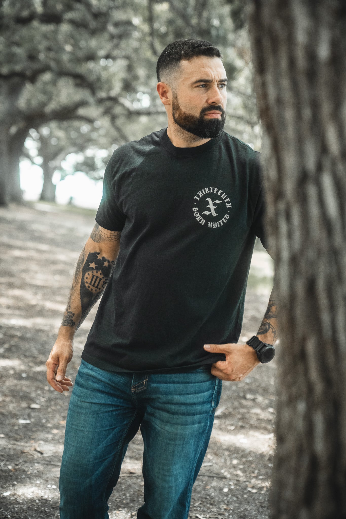 A bearded man with tattoos stands outdoors among trees, wearing the Born United Private Label’s Kingdom Protector Premium Tee and blue jeans, embodying the strength of Joshua 24:15—perfect inspiration for Christian home decor.