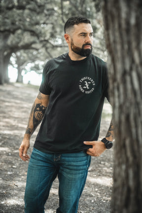 A bearded man with tattoos stands outdoors among trees, wearing the Born United Private Label’s Kingdom Protector Premium Tee and blue jeans, embodying the strength of Joshua 24:15—perfect inspiration for Christian home decor.