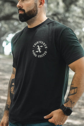 A bearded man wearing the black Kingdom Protector Premium Tee by Born United Private Label, with visible left arm tattoos, stands among trees—his look complements Christian home decor or Bible verse wall art such as Joshua 24:15.