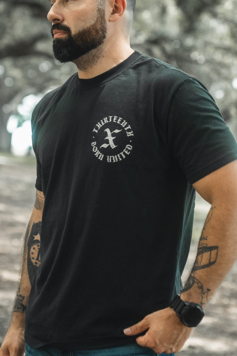 A bearded man wearing the black Kingdom Protector Premium Tee by Born United Private Label, with visible left arm tattoos, stands among trees—his look complements Christian home decor or Bible verse wall art such as Joshua 24:15.