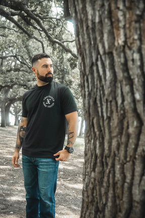 A man with short dark hair and a beard stands outdoors near a large tree, wearing the Born United Private Label Kingdom Protector Premium Tee, looking to his left as sunlight filters through the wooded area.