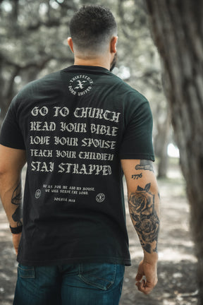 A tattooed man stands outdoors, seen from behind, wearing the Kingdom Protector Premium Tee by Born United Private Label. The black shirt features white text: GO TO CHURCH READ YOUR BIBLE LOVE YOUR SPOUSE TEACH YOUR CHILDREN STAY STRAPPED.
