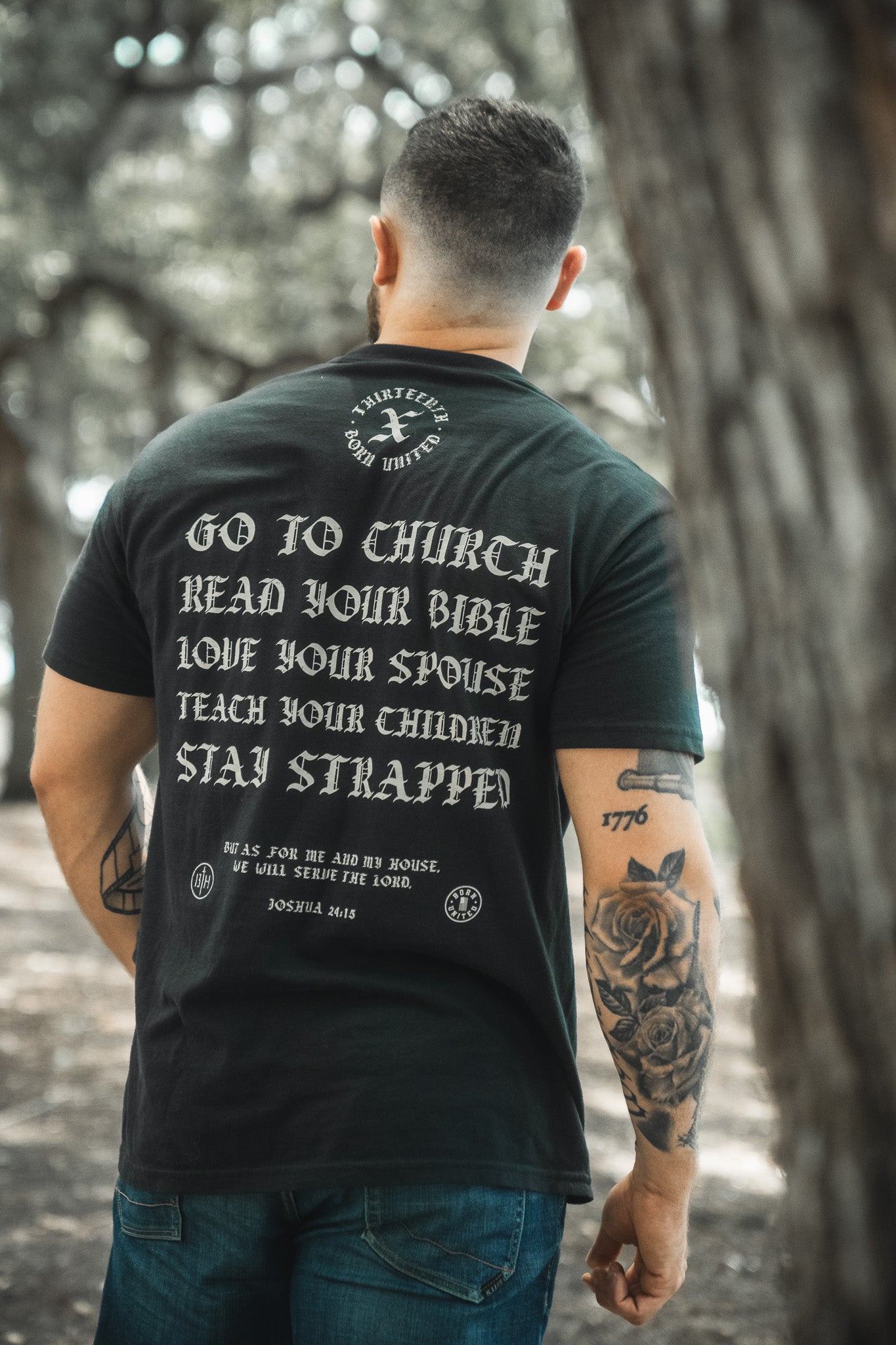 A tattooed man stands outdoors, seen from behind, wearing the Kingdom Protector Premium Tee by Born United Private Label, featuring white text inspired by Joshua 24:15: “Go to church, read your Bible, love your spouse, teach your children, stay strapped.”.