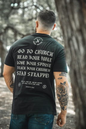 A tattooed man stands outdoors, seen from behind, wearing the Kingdom Protector Premium Tee by Born United Private Label, featuring white text inspired by Joshua 24:15: “Go to church, read your Bible, love your spouse, teach your children, stay strapped.”.