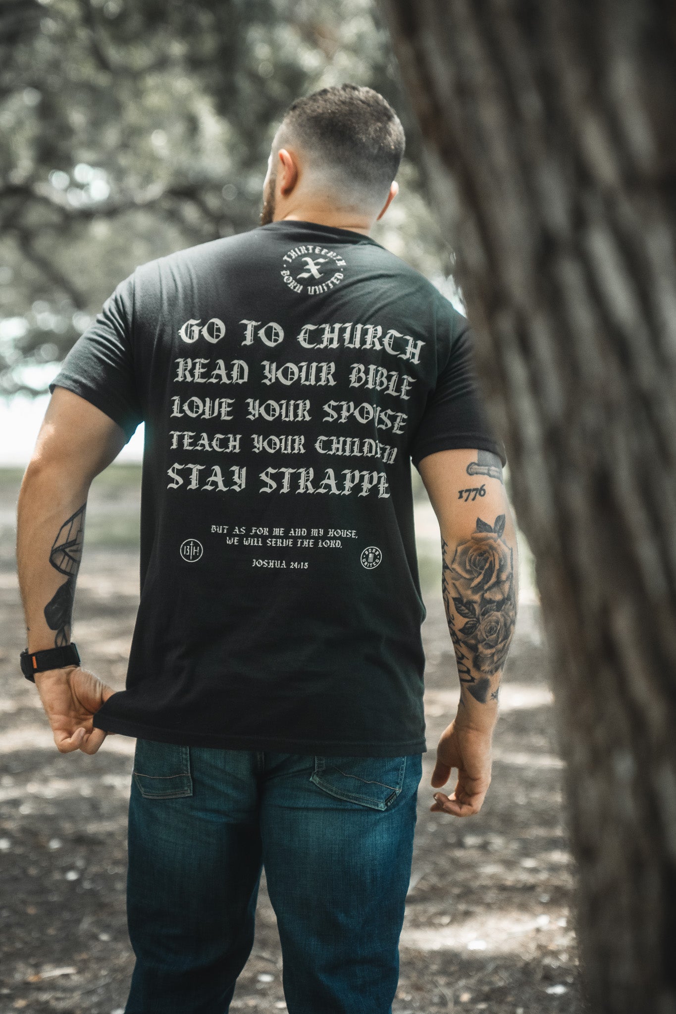 A man, seen from behind outdoors, wears the Born United Private Label "Kingdom Protector Premium Tee," a black shirt with the message: Go to church, read your Bible, love your spouse, teach your children, stay strapped. Arm tattoos visible.