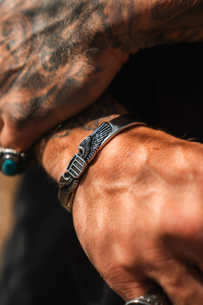 A close-up of a tattooed hand wearing the Relentless Betrayal Americana Bracelet—crafted from stainless steel with winged engravings, inspired by classic Americana designs—and a turquoise ring on another finger.