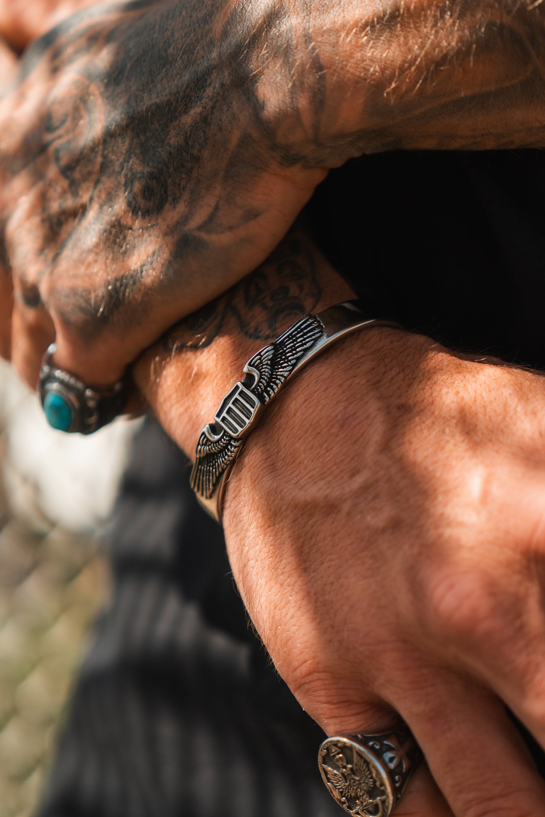A close-up of tattooed hands wearing silver rings and the Americana Bracelet by Relentless Betrayal—a stainless steel piece featuring an engraved winged design, proudly made in the USA.