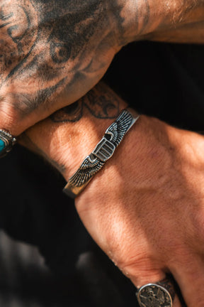A tattooed hand wearing the Relentless Betrayal Americana Bracelet, crafted in stainless steel with an engraved winged design and made in the USA, paired with a silver ring featuring an intricate pattern.