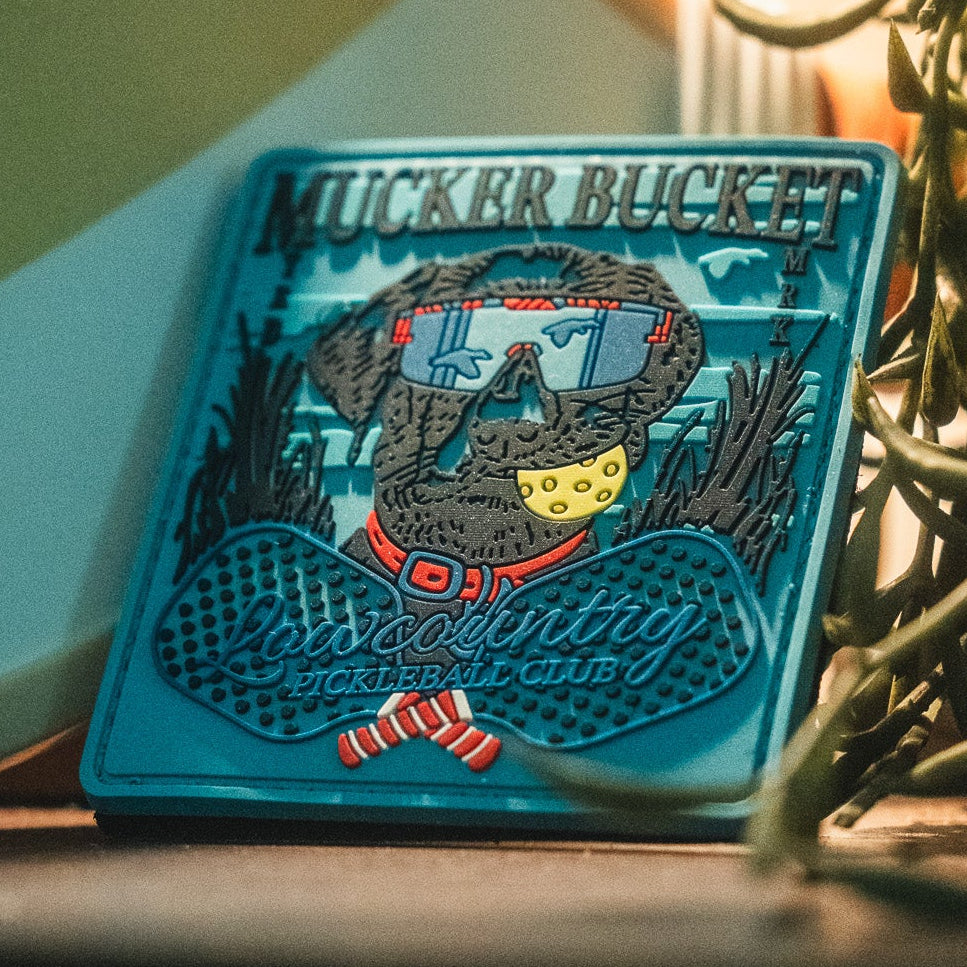 A vibrant 4x4 PVC patch from Mucker Bucket, the Pickleball Retriever Patch features a dog with sunglasses holding a pickleball, and attaches easily to any gear with Velcro for extra personality.