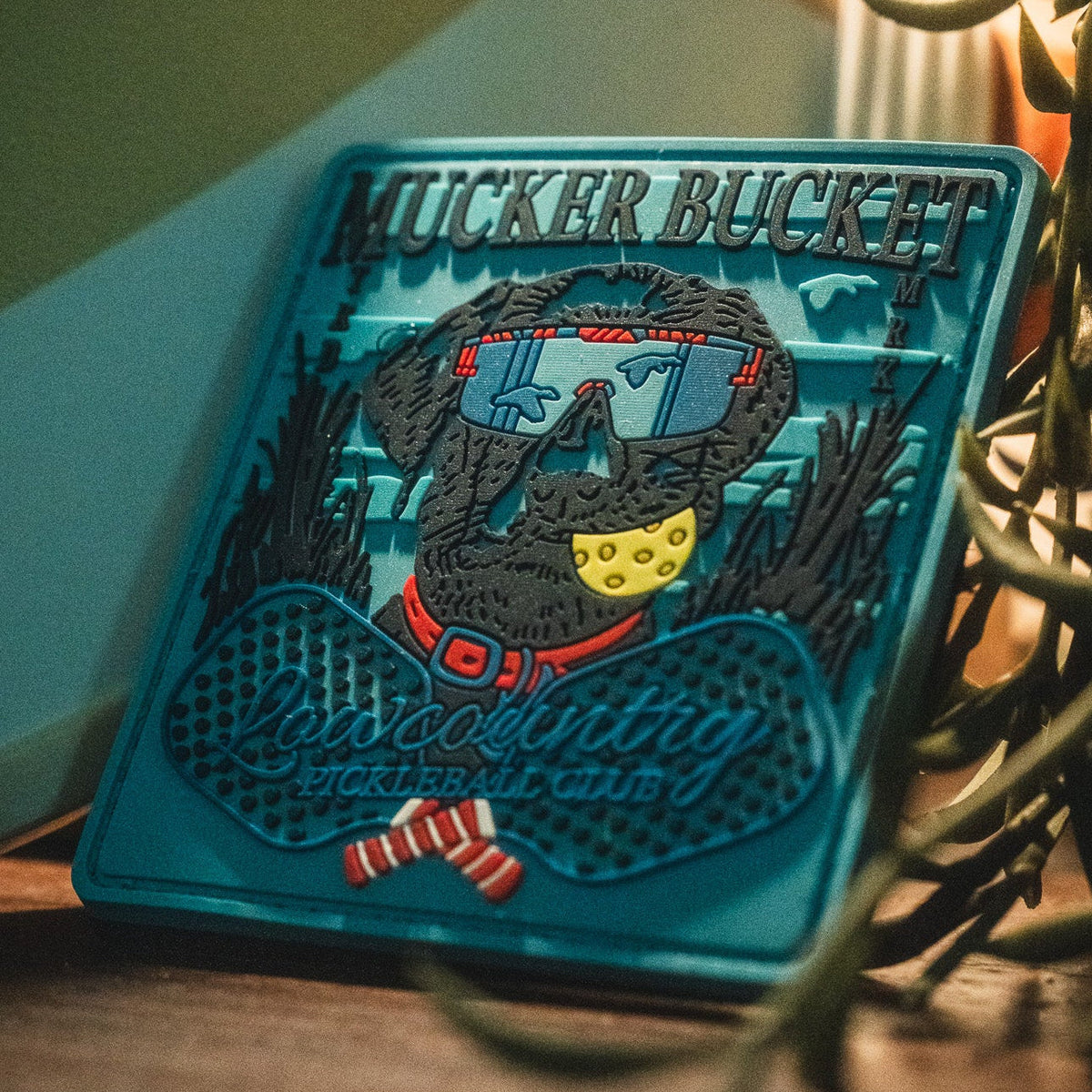 Introducing the Pickleball Retriever PVC Patch by Mucker Bucket—a vibrant 4x4 decorative patch featuring a dog sporting sunglasses and a bandana, along with the "Mucker Bucket" and "Lowcountry Pickleball Club" text. This stylish 3D PVC patch is both durable and eye-catching, making it the perfect accessory for your gear.