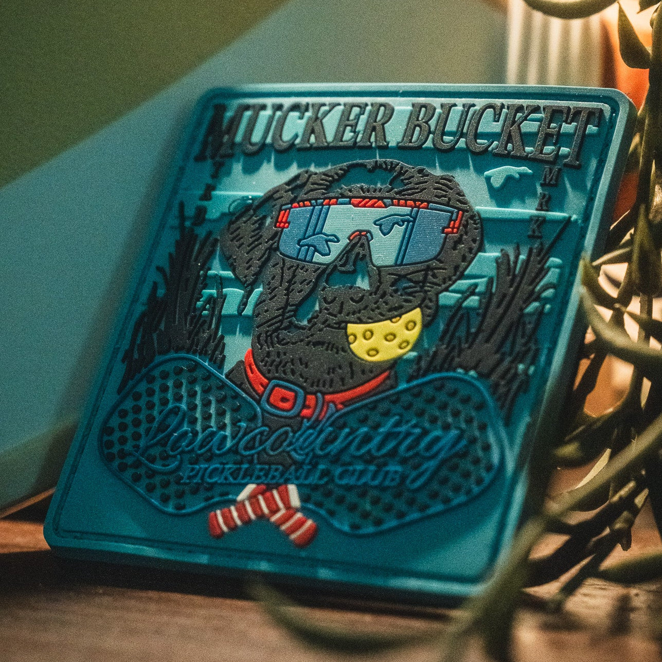 Introducing the Pickleball Retriever PVC Patch by Mucker Bucket—a vibrant 4x4 decorative patch featuring a dog sporting sunglasses and a bandana, along with the "Mucker Bucket" and "Lowcountry Pickleball Club" text. This stylish 3D PVC patch is both durable and eye-catching, making it the perfect accessory for your gear.