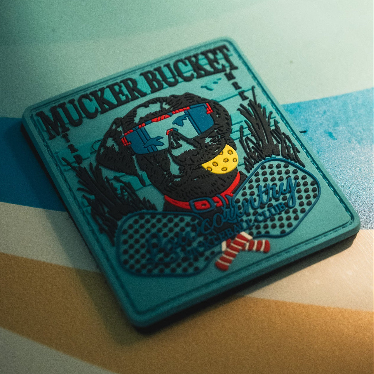 A 4x4 patch from Mucker Bucket features a bold rubber design of a dog wearing sunglasses, a red collar, and a yellow tag. The text "MUCKER BUCKET" appears at the top, with "Dog So Dirty, It’s Outrageous" below. This vibrant 3D PVC Pickleball Retriever Patch is both durable and eye-catching.