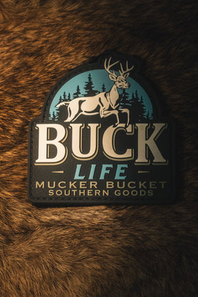 A rectangular patch with rounded top corners featuring a deer and pine trees on a teal background, with the text 'BUCK LIFE MUCKER BUCKET SOUTHERN GOODS' in cream and dark lettering, placed on a brown fur surface.