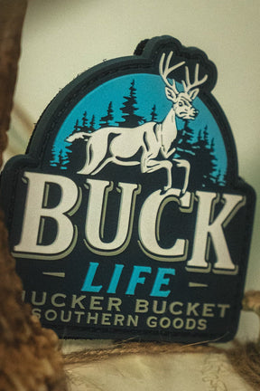 The Buck Life PVC Patch by Mucker Bucket showcases a white deer and trees logo, framed by bold antlers and labeled with "Mucker Bucket Southern Goods," expertly crafted as a durable 4x4 Velcro patch for your favorite gear.