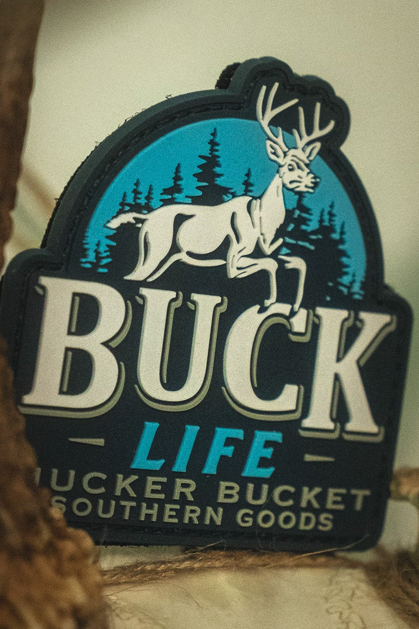 The Buck Life PVC Patch by Mucker Bucket showcases a white deer and trees logo, framed by bold antlers and labeled with "Mucker Bucket Southern Goods," expertly crafted as a durable 4x4 Velcro patch for your favorite gear.