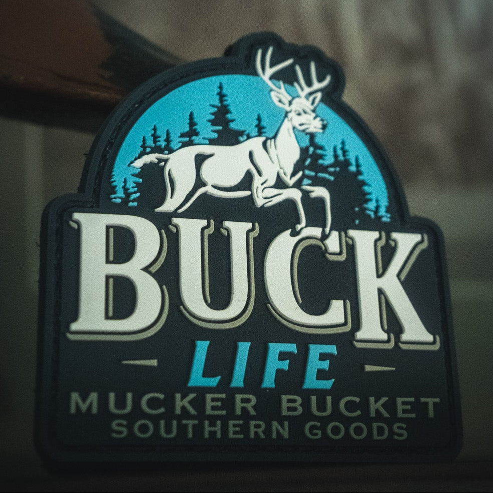 Sticker with a white deer and pine trees reads: Buck Life by Mucker Bucket, perfect for matching with your favorite 4x4 or adding to your Velcro patch collection. Features the bold Buck Life PVC Patch from Mucker Bucket Southern Goods.