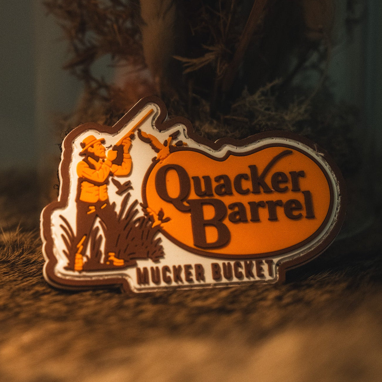 A Quacker Barrel PVC Patch by Mucker Bucket, featuring a person with birds and shaped like a state outline, rests on a textured brown surface—showcasing the bold style characteristic of 3D PVC patches.