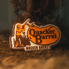 Introducing the Quacker Barrel PVC Patch by Mucker Bucket—a badge shaped like Kentucky, featuring the text "Quacker Barrel Mucker Bucket" and an illustration of a man aiming a gun in an orange and brown color scheme. This unique 3D PVC patch measures 4x4 inches and comes with Velcro backing for easy attachment.