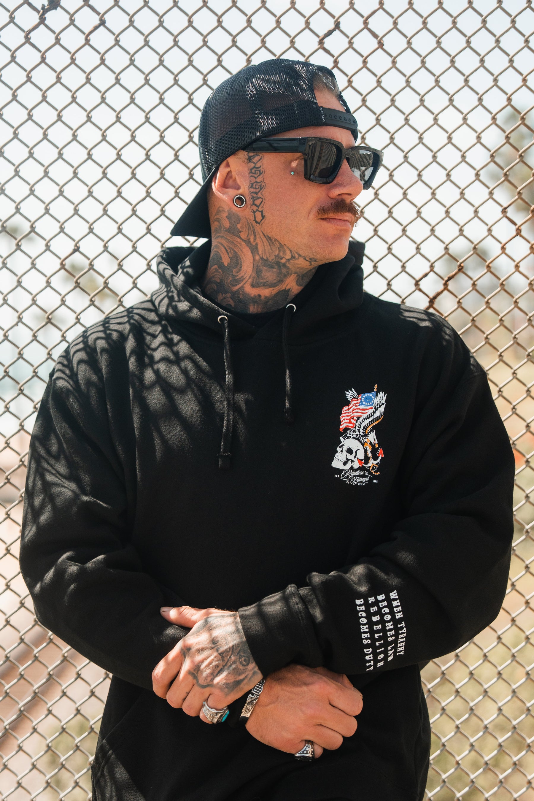 A tattooed man in the Relentless Betrayal Revolution Premium Hoodie, featuring a dragon and skull design, sunglasses, and a black backward cap, stands with arms crossed in front of a chain-link fence.