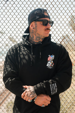 A tattooed man in the Relentless Betrayal Revolution Premium Hoodie, featuring a dragon and skull design, sunglasses, and a black backward cap, stands with arms crossed in front of a chain-link fence.