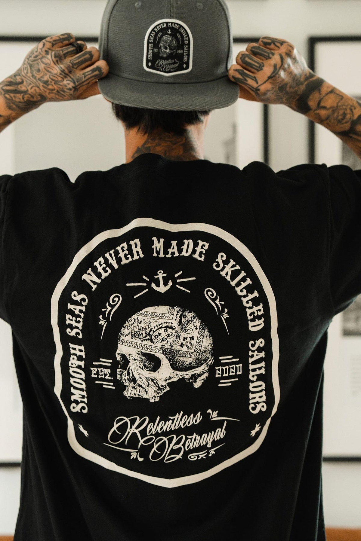 Tattooed arms visible, a resilient man dons the Skilled Sailor Tee by Relentless Betrayal, showcasing a large skull and nautical design with the words Smooth seas never made skilled sailors. Adjusting his gray cap, he embodies determination while confidently facing away from the camera.