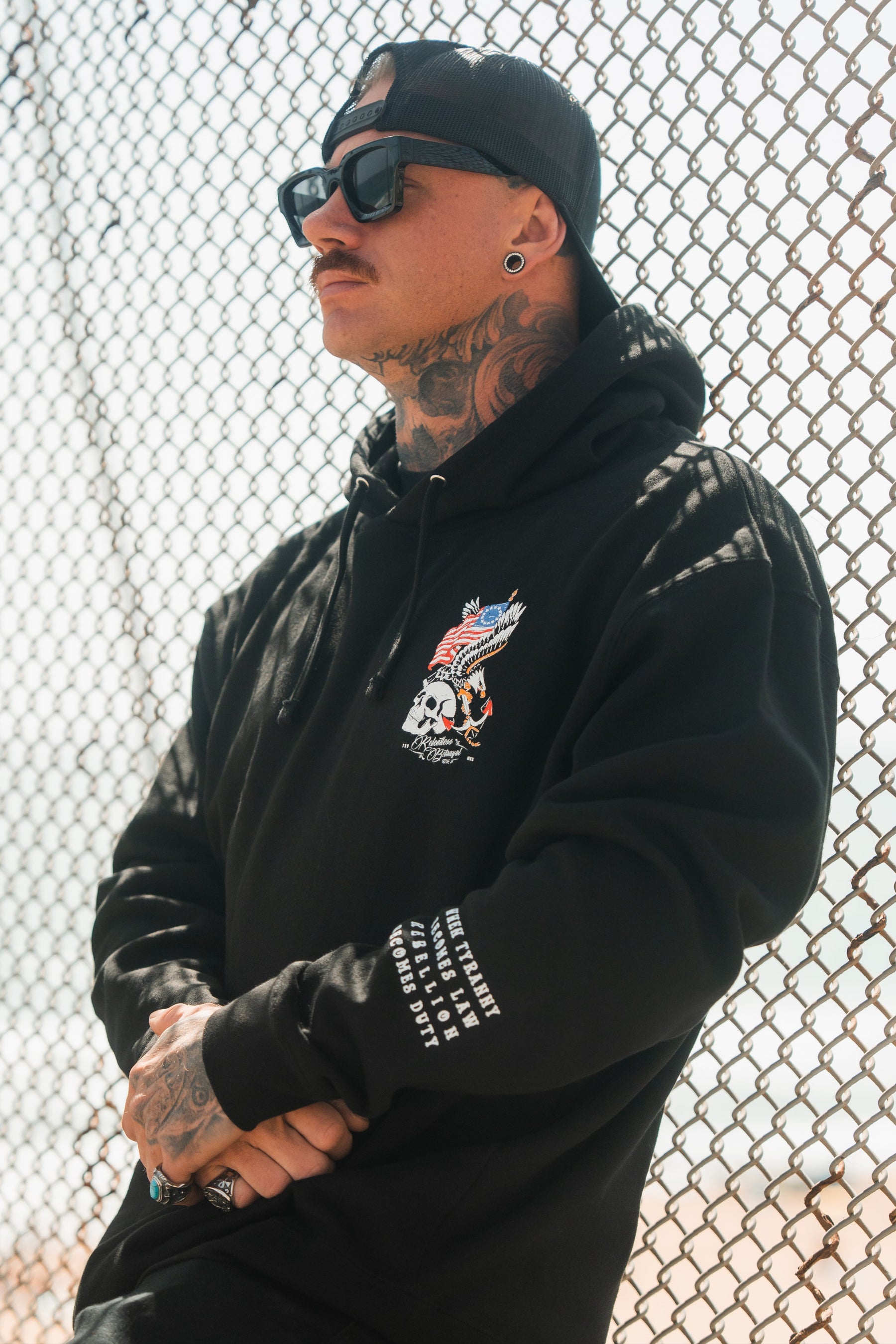 A tattooed man in sunglasses and a backwards cap wears the Relentless Betrayal Revolution Premium Hoodie with a bold skull and eagle graphic, standing before a chain-link fence on a sunny day.