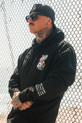 A tattooed man in sunglasses and a backwards cap wears the Relentless Betrayal Revolution Premium Hoodie with a bold skull and eagle graphic, standing before a chain-link fence on a sunny day.