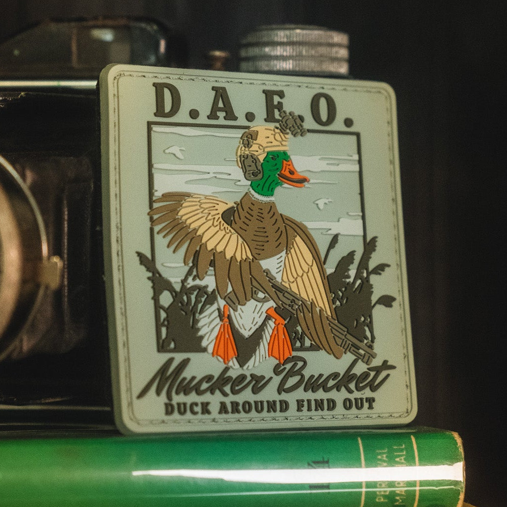Showcasing a duck in colonial attire holding a musket with the text "Duck Around & Find Out" and "Mucker Bucket" against a scenic nature background, the Mucker Bucket Duck Around & Find Out PVC Patch is a detailed 4x4 inch 3D PVC Velcro patch—perfect for boldly expressing your personality on any gear.