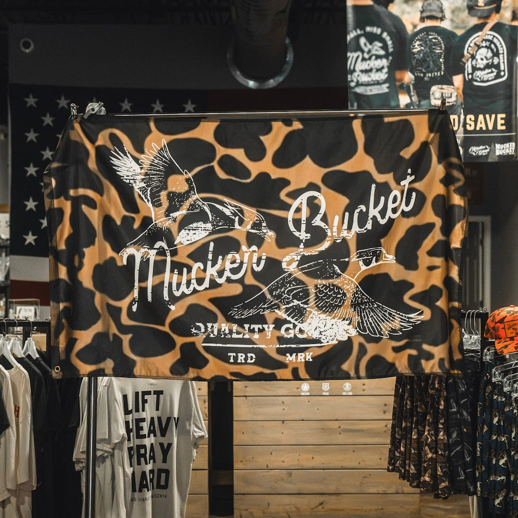 A Wood Ducks Duck Camo Flag from Mucker Bucket, featuring a camouflage pattern with flying duck illustrations, sturdy metal grommets, and durable polyester construction, hangs inside a clothing store.