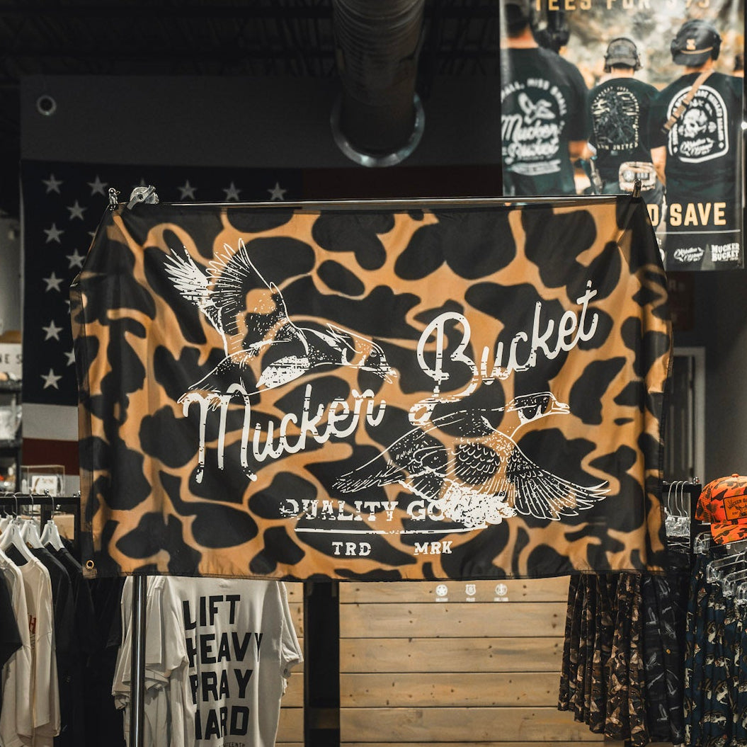 A Wood Ducks Duck Camo Flag by Mucker Bucket, crafted from durable polyester and featuring the Mucker Bucket Quality Goods TRD MRK logo along with two flying ducks, is displayed in a clothing store above racks of shirts. Metal grommets provide easy hanging and lasting strength.