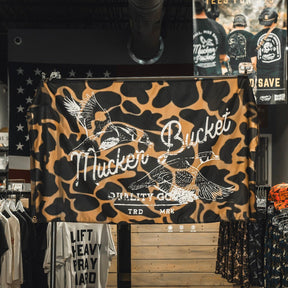 A Wood Ducks Duck Camo Flag by Mucker Bucket, crafted from durable polyester and featuring the Mucker Bucket Quality Goods TRD MRK logo along with two flying ducks, is displayed in a clothing store above racks of shirts. Metal grommets provide easy hanging and lasting strength.