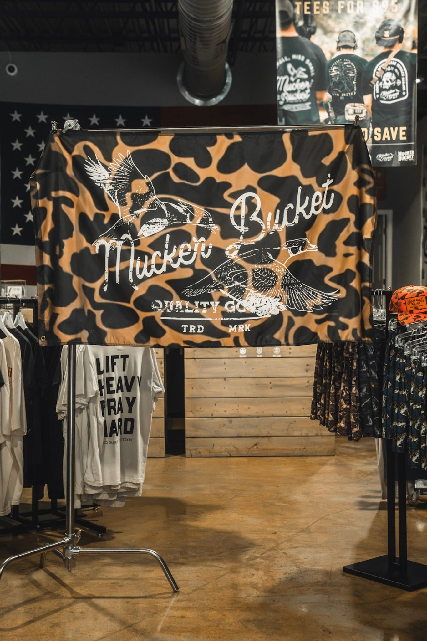 Wood Ducks Duck Camo Flag by Mucker Bucket displayed in a clothing store surrounded by shirts and apparel, featuring flying bird graphics and crafted from durable polyester with metal grommets for easy hanging.