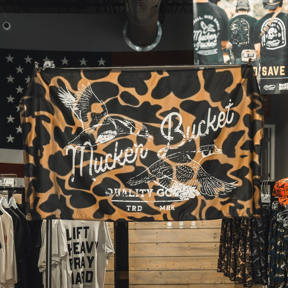 A Wood Ducks Duck Camo Flag by Mucker Bucket, featuring a camouflage pattern with flying ducks, is displayed in a clothing store. The flag is made from durable polyester with sturdy metal grommets. Racks of apparel and another Stacking Ducks Flag can be seen in the background.