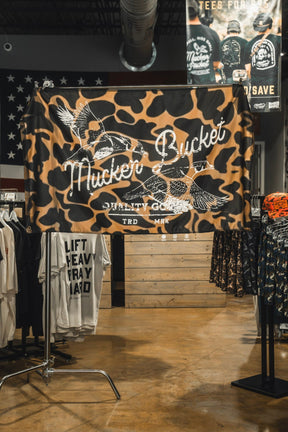 Wood Ducks Duck Camo Flag by Mucker Bucket displayed in a clothing store surrounded by shirts and apparel, featuring flying bird graphics and crafted from durable polyester with metal grommets for easy hanging.