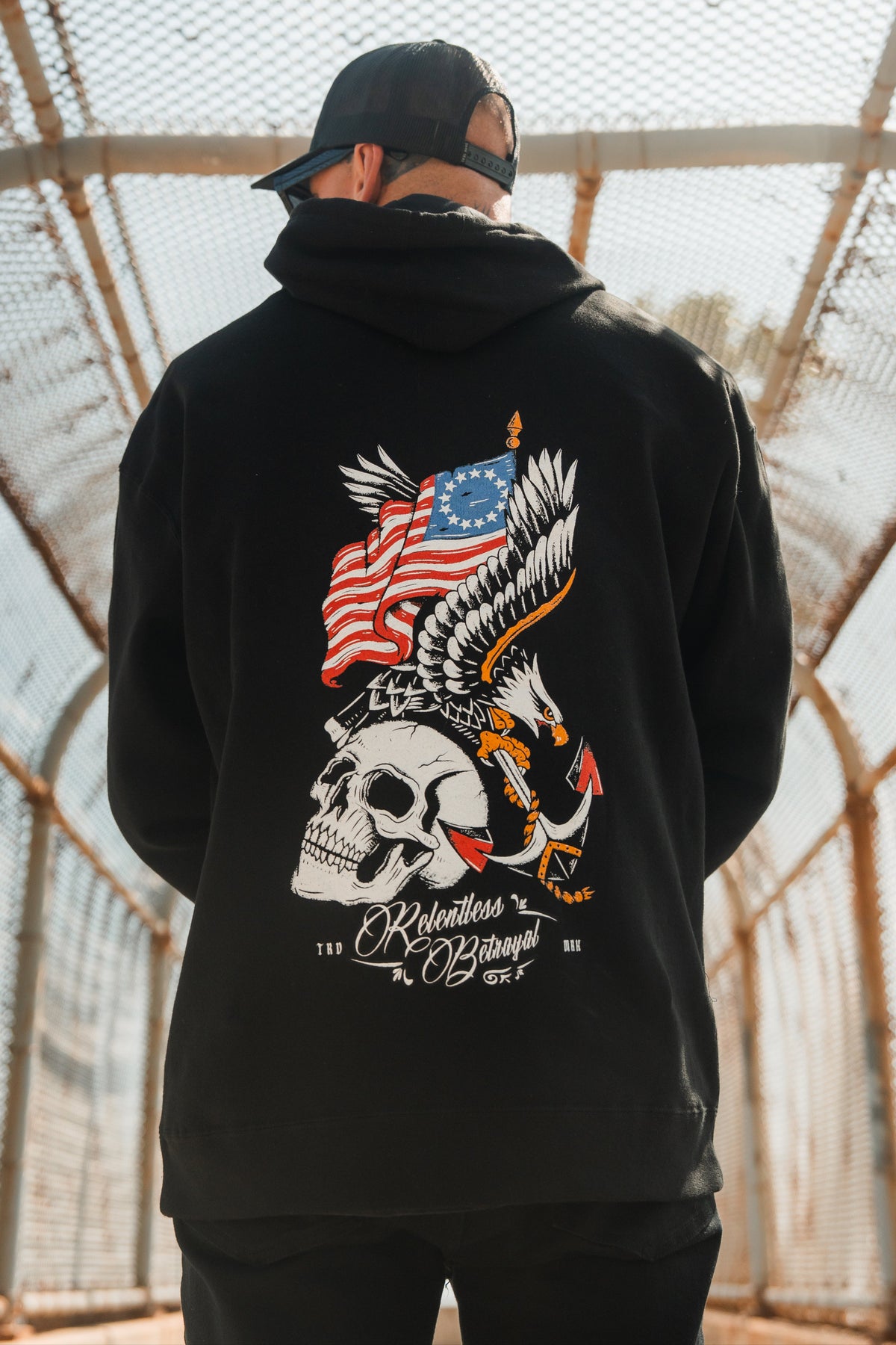 A man stands on a fenced pedestrian bridge, facing away from the camera, wearing the Revolution Hoodie by Relentless Betrayal. This premium black hoodie features an eagle, skull, American flag, and anchor graphic—delivering integrity and bold style in every detail.