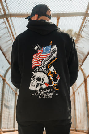 A person wearing the Relentless Betrayal Revolution Premium Hoodie—with a bold skull, eagle, American flag, text details, and an anchor for integrity—stands on a fenced bridge while sporting a backward black cap.