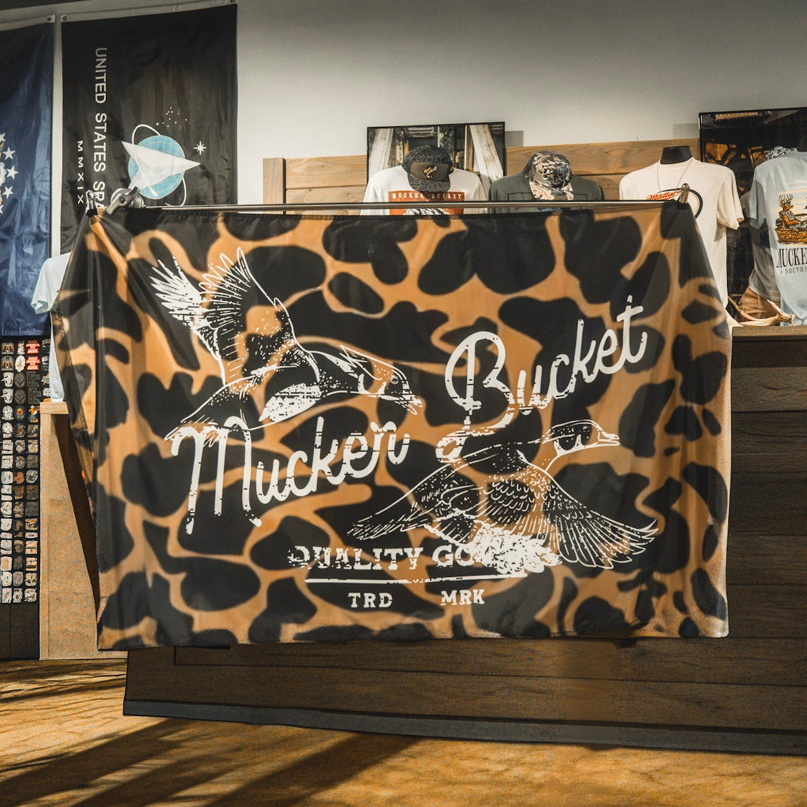 A durable polyester “Wood Ducks Duck Camo Flag” by Mucker Bucket, showcasing a brown and black camouflage design with two flying ducks and the phrase “Nucker Bucket Quality Goods TRD MRK,” displayed inside a retail shop.