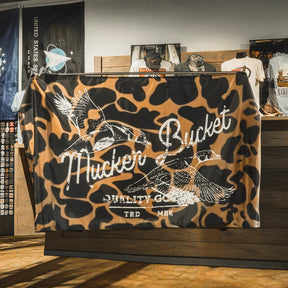 A durable polyester “Wood Ducks Duck Camo Flag” by Mucker Bucket, showcasing a brown and black camouflage design with two flying ducks and the phrase “Nucker Bucket Quality Goods TRD MRK,” displayed inside a retail shop.