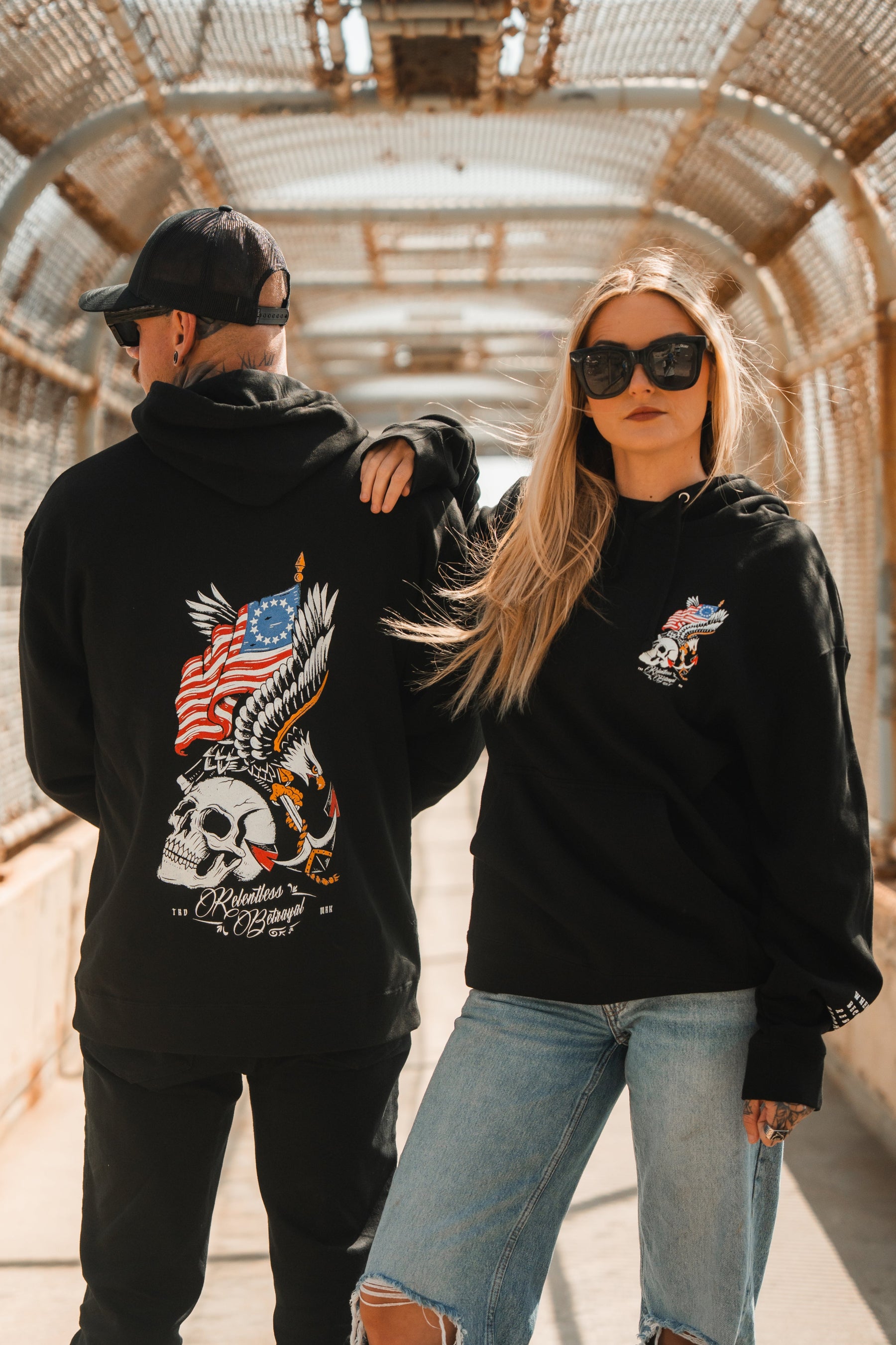 A man and woman stand on a pedestrian bridge in Relentless Betrayal Revolution Premium Hoodies. The man's features an eagle, skull, and American flag; the confident woman pairs hers with sunglasses and ripped jeans, hand on his shoulder.