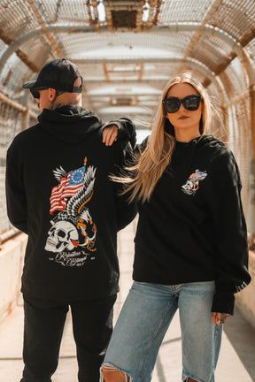 A man and woman stand on a pedestrian bridge in Relentless Betrayal Revolution Premium Hoodies. The man's features an eagle, skull, and American flag; the confident woman pairs hers with sunglasses and ripped jeans, hand on his shoulder.