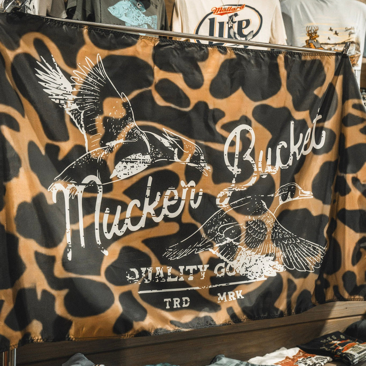 The Wood Ducks Duck Camo Flag by Mucker Bucket features a brown and black duck camo pattern with two flying ducks and the text “Mucker Bucket Quality Goods TRD MRK” printed on it. Made from durable polyester, it includes metal grommets for easy hanging.