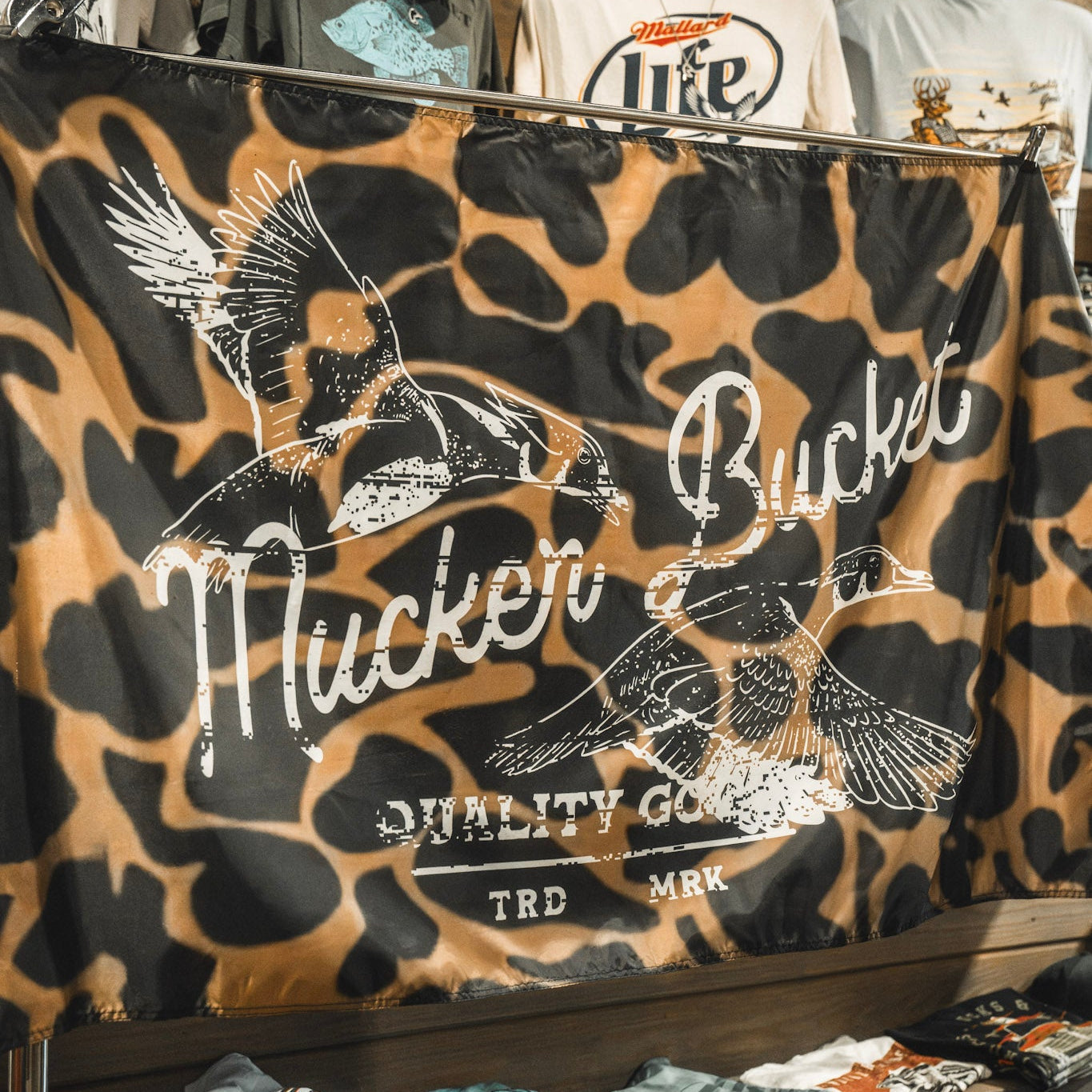 The Wood Ducks Duck Camo Flag by Mucker Bucket features a brown and black duck camo pattern with two flying ducks and the text “Mucker Bucket Quality Goods TRD MRK” printed on it. Made from durable polyester, it includes metal grommets for easy hanging.